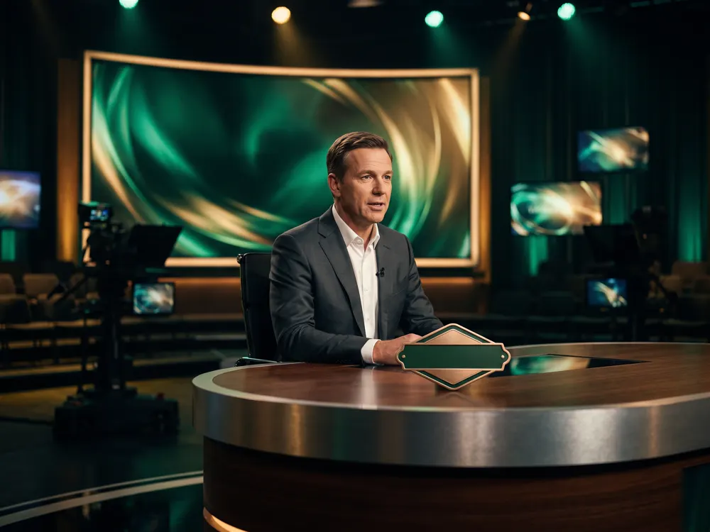 A presenter at a broadcast desk in front of a large screen